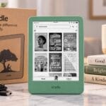 Matcha green Amazon Kindle 16GB showing its library screen, standing beside its kraft box, a USB-C cable, a speckled mug and a stack of books.