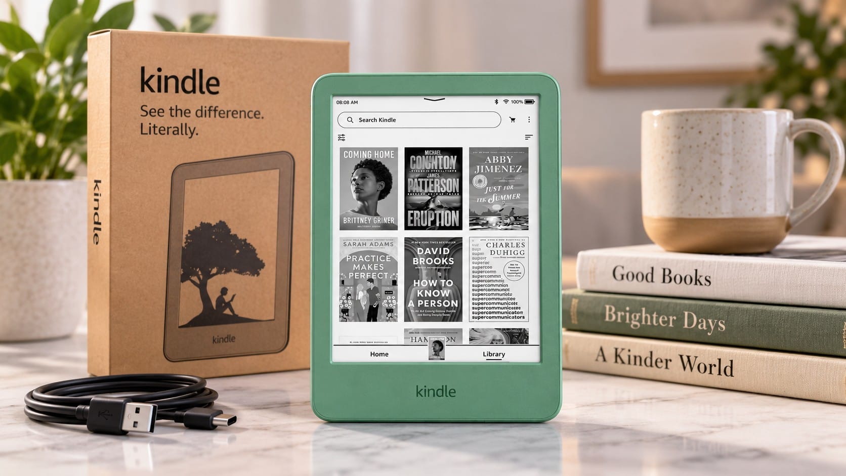 Matcha green Amazon Kindle 16GB showing its library screen, standing beside its kraft box, a USB-C cable, a speckled mug and a stack of books.