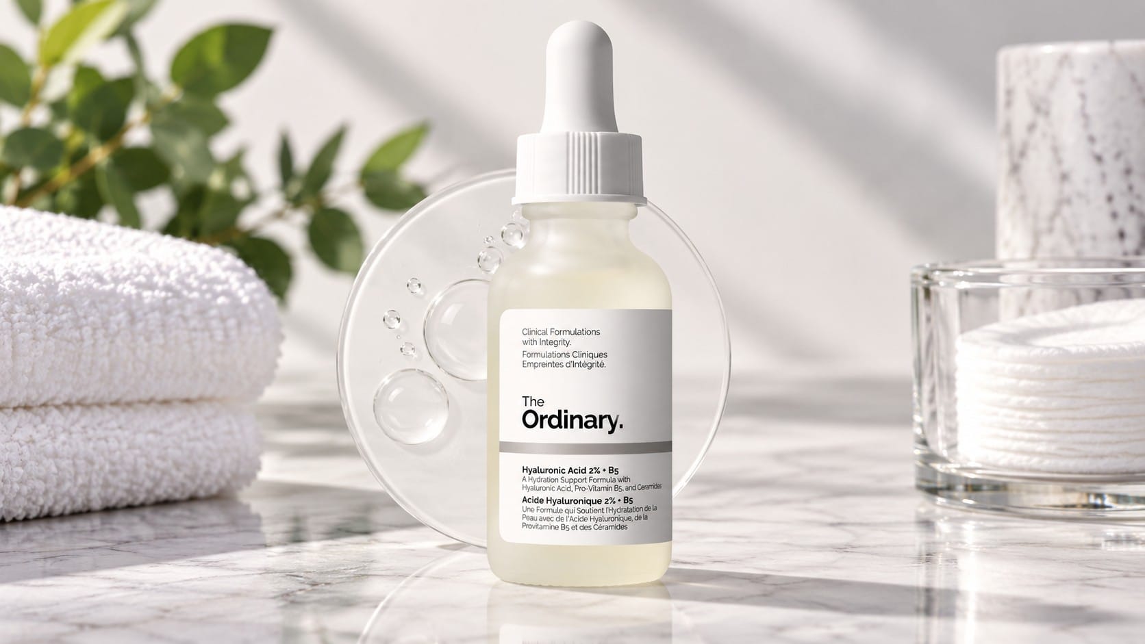 The Ordinary Hyaluronic Acid 2% + B5 dropper bottle on a marble bathroom counter in front of a clear serum swatch, with white towels, a plant and cotton pads.