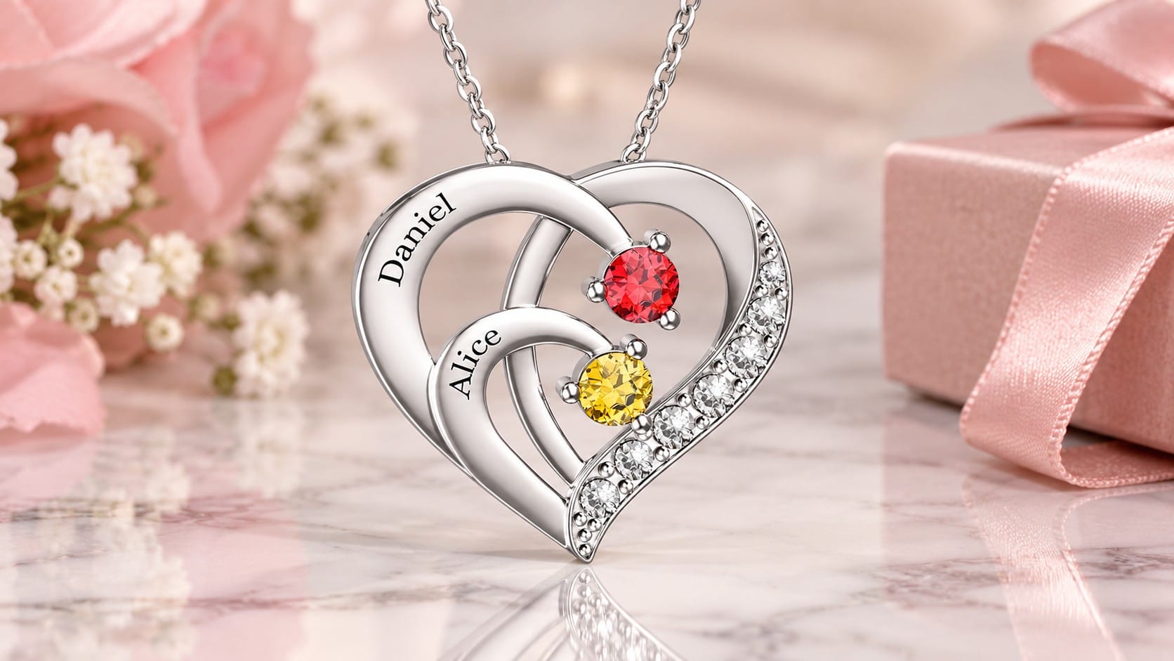 Silver heart pendant engraved with the names Daniel and Alice, set with red and yellow simulated birthstones and a row of crystals, on a blush marble surface with roses and a gift box.